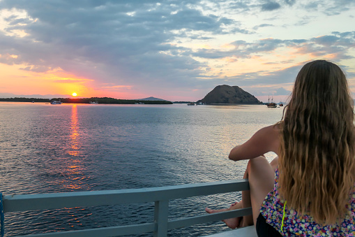 Girl with long hair on a small boat enjoying beautiful sunset in Komodo National Park, Flores, Indonesia. Sun sets over the horizon line. Girl is admiring the natural spectacle. Happiness and freedom