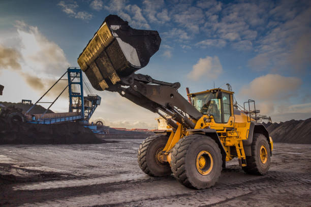 양동이 로더는 석탄의 검은 산을 배경으로 열린 항구 창고에서 석탄을 적재합니다. - construction equipment industrial equipment loading construction 뉴스 사진 이미지