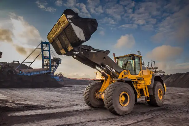 Photo of A bucket loader carries out loading of coal in an open port warehouse on a background of black mountains of coal