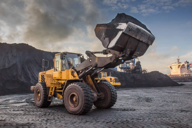 un caricatore a secchio effettua il carico di carbone in un magazzino portuale aperto su uno sfondo di montagne nere di carbone - loading wheel mining equipment foto e immagini stock