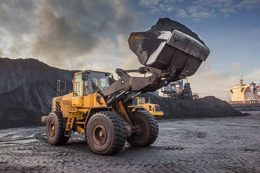 A bucket loader carries out loading of coal in an open port warehouse on a background of black mountains of coal