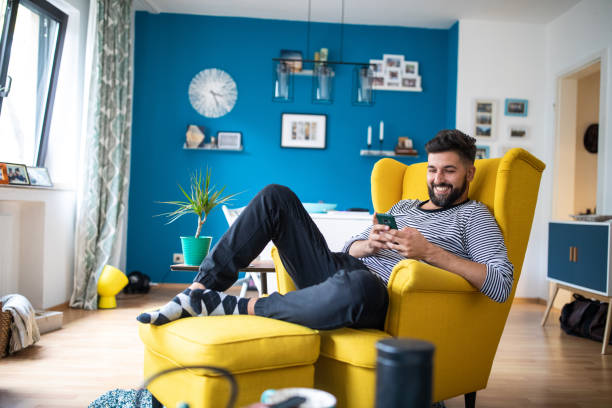 Happy bearded man sitting on armchair and using smart phone Young bearded mid adult man sitting in yellow armchair at apartment and using mobile phone and smiling macho photos stock pictures, royalty-free photos & images