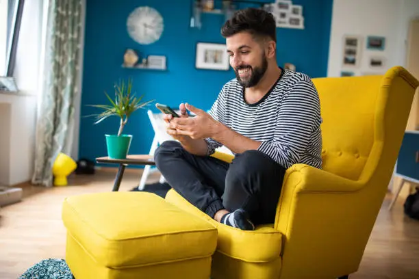 Photo of Happy bearded man sitting in apartment and sending text messages