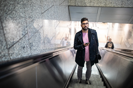Businessman using escalator in subway, holding mobile phone and looking around