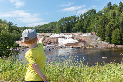 A little redhead boy is looking at a waterfall in summer. He is pointing the waterfall and river to his parents. It is a beautiful sunny day. Location : Parc de la Chûte de Sault-Au-Mouton, Quebec, Canada