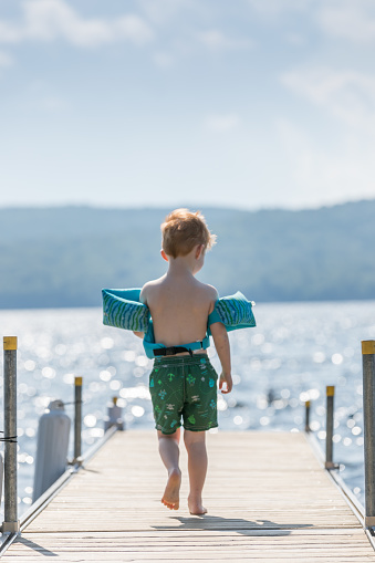 Little redhead boy walking on a pier in summer. He is wearing is life jacket to go swimming. It is a beautiful summer day and the sunlight is reflecting on the lake. There is a mountain range in the background.