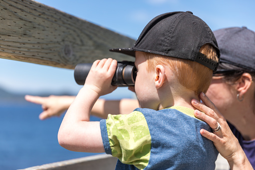 A little redhead boy and his mother are whale watching and looking at the view through binoculars in summer. The mother is pointing a whale passing by. Location : Baie Sainte-Marguerite, Lac Saint-Jean, Canada.