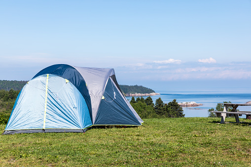 A blue tent is located on a mountain peek during summer. The camping spot offers a beautiful view of the river. It is located in Cote nord, Les Bergeronnes, Quebec, Canada.