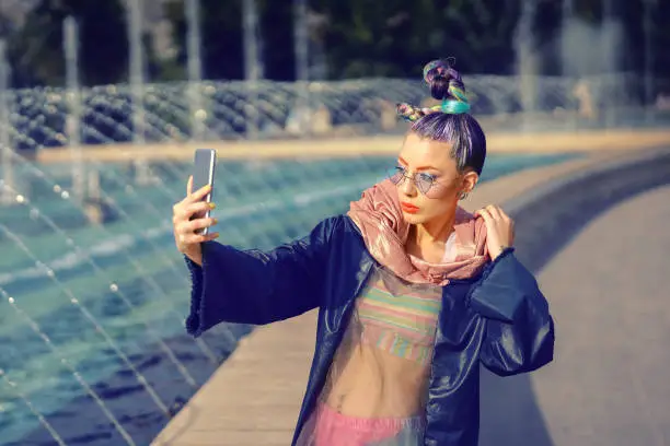 Cool funky hipster young fashion influencer girl with crazy hair and avant garde style taking selfie on street - woman sharing trendy content on streaming platform and social media platform