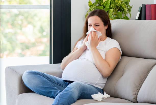 ill pregnant woman coughing sitting on a couch Shot of a ill pregnant woman coughing sitting on a couch at home sneezeweed stock pictures, royalty-free photos & images