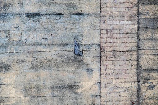 A section of an old, damaged concrete wall with a vertical column of bricks shows a variety of textures and lines where it has been patched and repaired with plaster, concrete and a piece of wood.