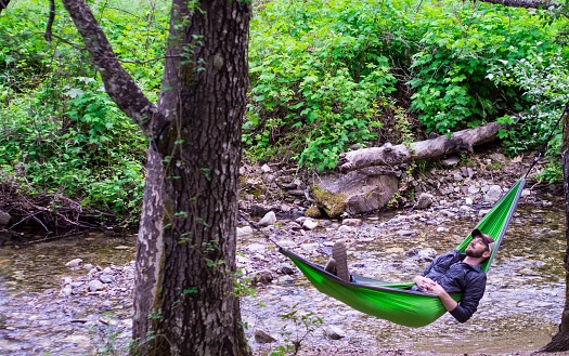 A man takes a nap in a hammock next to a crystal clear stream.