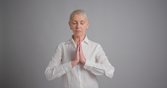 Peaceful elderly woman practicing yoga and meditation. Calm woman making Namaste gesture on gray background.