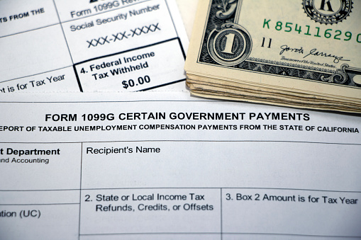 Closeup of overlapping tax forms, Form 1099G Certain Government Payments, Federal Income Tax Withheld $0.00, with stack of cash, blank form boxes.