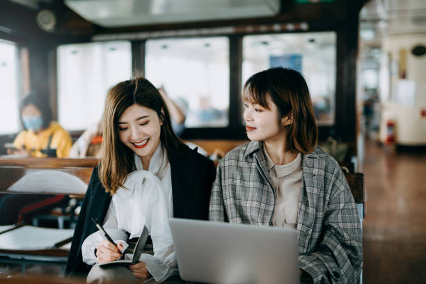 istock Two Asian young businesswomen using laptop and discussing a project while riding on a ferry 1202006351