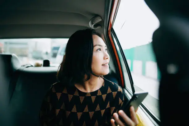 Photo of Asian woman sitting in the back of a taxi looking out the window and smiling while commuting in the city