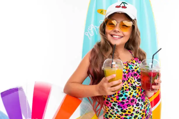 Photo of Girl with long crucified hair in sunglasses and swimsuit stands near inflatable mattresses with fresh fruit cocktails and smiles isolated on a white background