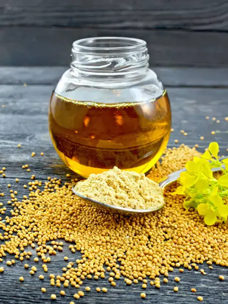 Mustard powder in a spoon, oil in a glass jar, seeds and mustard flower against a black wooden board