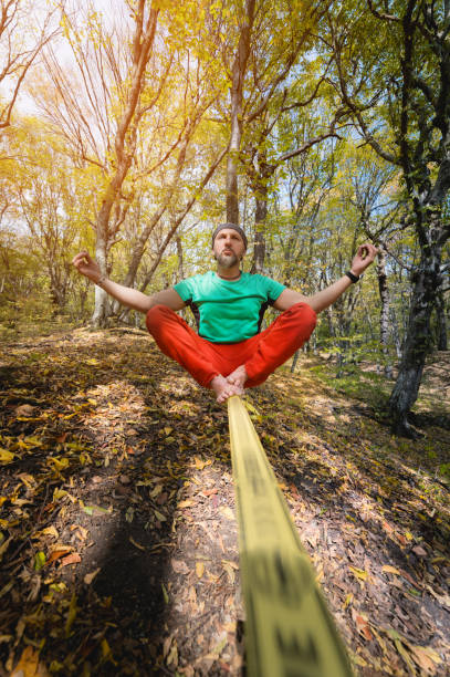 istock Wide angle male tightrope walker balancing while meditation sitting barefoot on slackline in autumn forest. The concept of outdoor sports and active life of people aged 1202032495