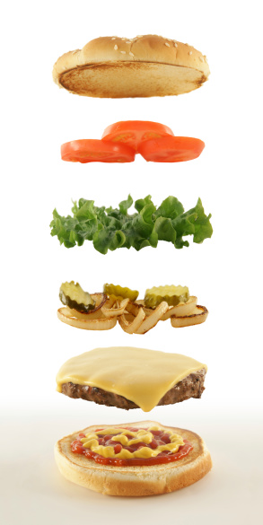 High resolution, digital capture of an exploded view of a classic cheeseburger, showing individual ingredients as floating layers, with central perspective convergence. Cheeseburger consists of a toasted top and bottom bun, catsup and mustard, hamburger patty with melted cheese, grilled onions and pickles, lettuce, and tomatoes. Shot against a pure white background on a slightly gray surface.
