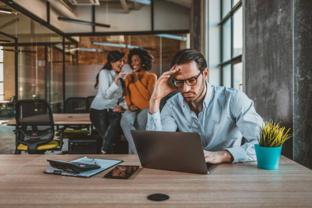 Positive multi-ethnic employees gossiping about tired male colleague Lonely and sad businessman at office of company. Photo of Sad businessman with female team in the background. Male office manager unable to cope with too many tasks from his boss and supervisor, suffering stress, pressing her head with desperate expression. harassment stock pictures, royalty-free photos & images