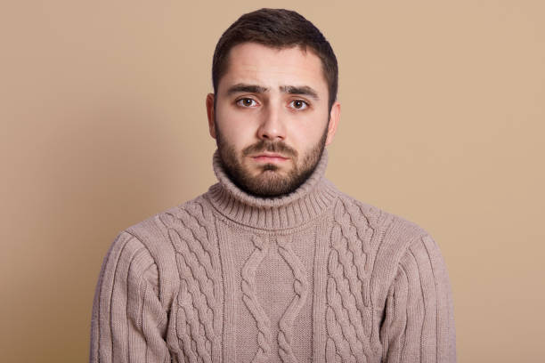 istock Horizontal shot of attractive young man with beard posing in studio against beige wall, looking at camera with serious facial expression, wearing knitted sweater, being in bad mood, having problems. 1202046551
