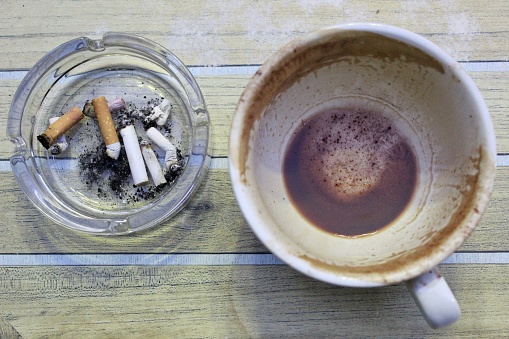 Empty Cup of Coffee and Cigarette Ends in Clear Glass Ashtray on Wooden Table Background
