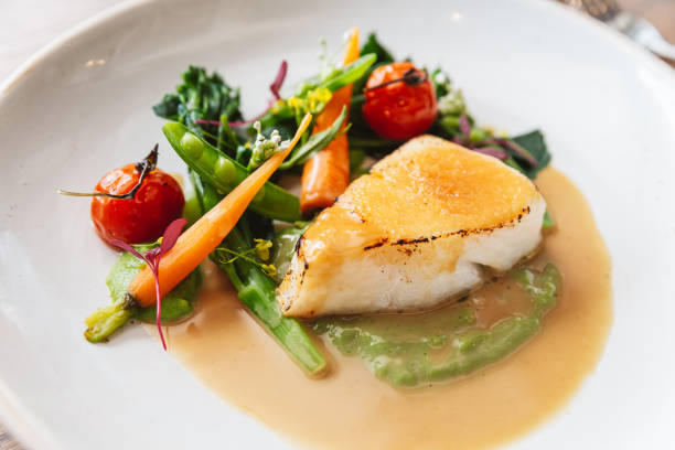 istock Close-up Pan seared Cod fish fillet with tomato, bean and carrot in gravy sauce. Served in a white plate with a silver knife and fork. 1202064700