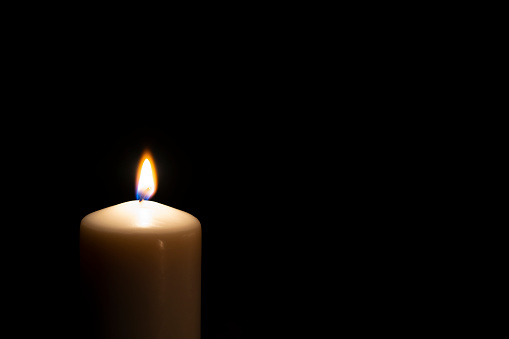 Single lit candle with quite flame in the dark. The composition is at the left of an horizontal frame leaving useful copy space for text and/or logo at the right. High resolution 42Mp studio digital capture taken with Sony A7rii and Sony FE 90mm f2.8 macro G OSS lens