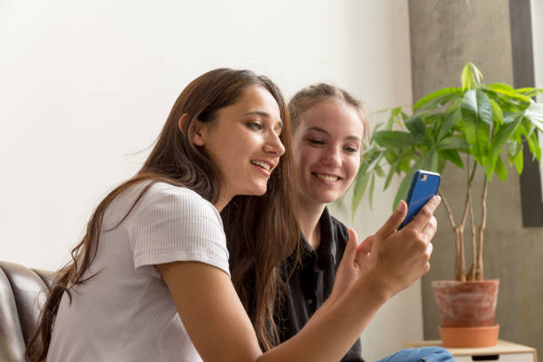 Two Young Women Seated Side by Side Looking Happily at Smartphone