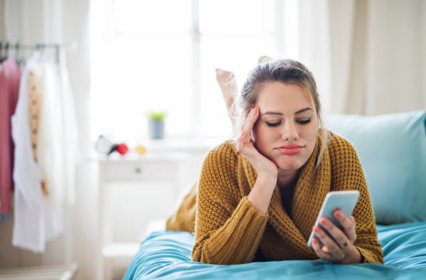 bored young woman with smartphone lying on bed indoors at home. - dull colors imagens e fotografias de stock