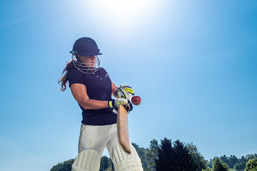 Low angle photo of a female cricket batter hitting the ball with the sun and sky in the background.