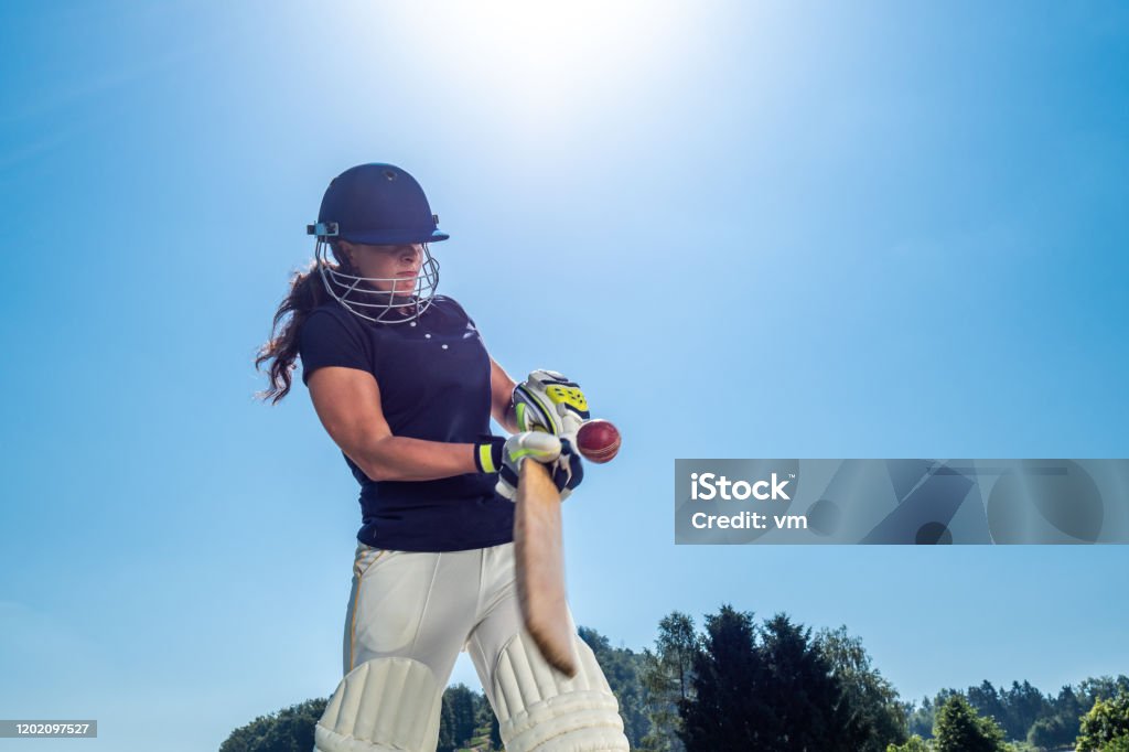 Female cricket batter hitting the ball Low angle photo of a female cricket batter hitting the ball with the sun and sky in the background. Sport of Cricket Stock Photo
