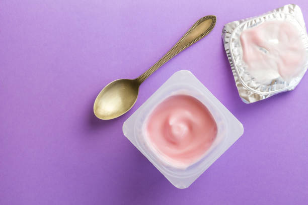 yogur con sabor a fruta de fresa saludable con coloración natural en taza de plástico aislada sobre fondo púrpura con cuchara pequeña y tapa de papel de aluminio - yogurt yogurt container strawberry spoon fotografías e imágenes de stock