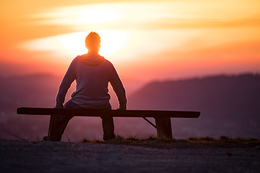 Adult Man Sitting on a Bench Alone and Watching Sunset Above a Landscape.