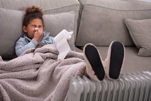 Sick child blowing nose into a toilet paper, covered with a blanket, wearing slippers, heating up feet on an oil radiator heater. The kid with flu cold or allergy symptoms sitting on a couch at home in the winter season.