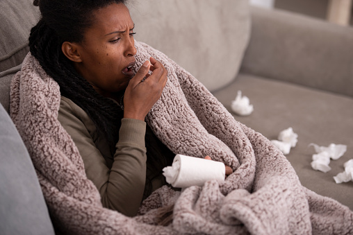 Sick woman with her hand over her mouth coughing, sitting wrapped with a warm blanket on the sofa at home.  Female with cold or allergies.