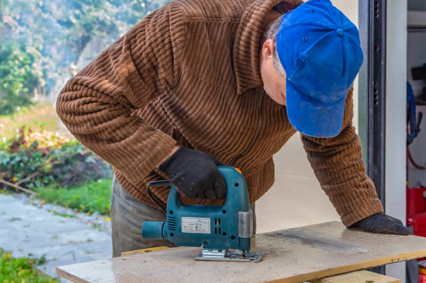 istock A man in the black working gloves and a brown jacket and a blue hat cuts a board using a Jigsaw power tool on a work bench 1202107729
