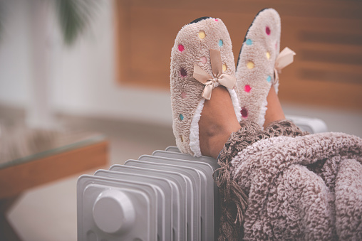 Little girl's feet in slippers with Polka dots and ribbon bow, lying on radiator heater in the living room. Legs covered with a warm blanket. Kid staying at home in the winter season.