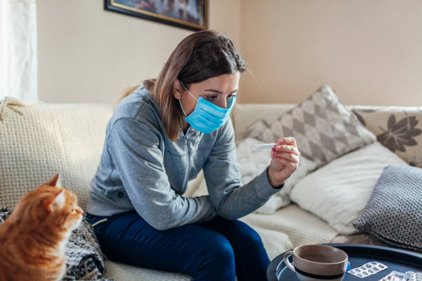 Ill woman checking thermometer having fever. Girl wearing protective mask while having cold, flu at home. Healthcare Ill infected woman checking thermometer having fever. Girl wearing protective mask while having cold, flu taking pills at home. Healthcare covid thermometer stock pictures, royalty-free photos & images