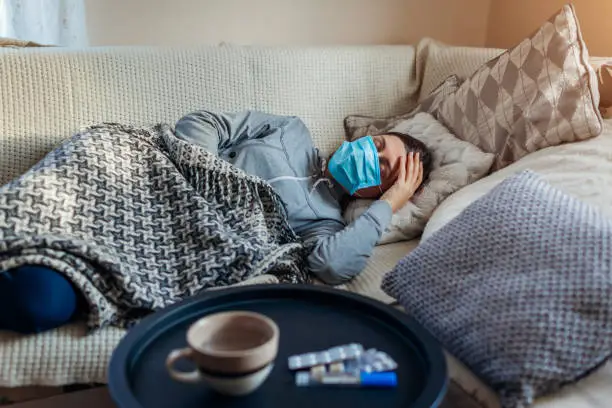 Sick woman having flu or cold. Girl lying in bed having headache wearing protective mask by pills and water on table.