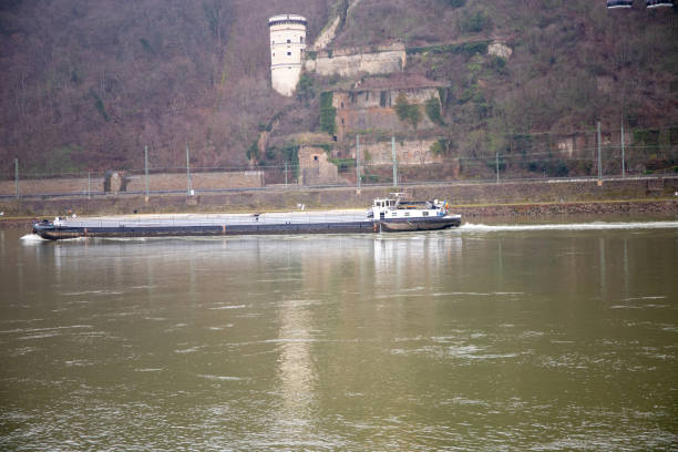 Painted red, white and black, a barge on the Rhine River in Germany carrying economic cargo, moves upriver near Bacharat in Rhineland-Palatinate. The area is part of the Rhine Gorge, which was added to the UNESCO list of World Heritage Sites in June 2002. With a red navigation bouy seen in the flowing water, this inland vessel helps to facilitate commerce in Germany and, by extension, the world economy. A road and steep valley side with lush foliage are seen behind the river shore line.