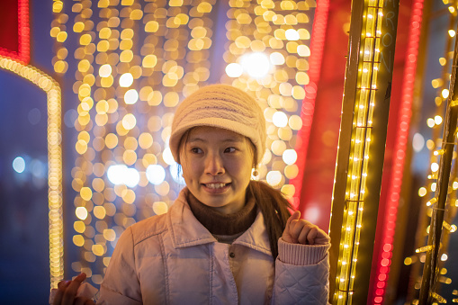 Young woman from Chine visiting Europe and walking on square in city center. . She is alone and happy when posing with street Christmas decoration
