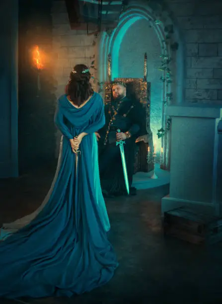 Photo of Luxury beauty Queen turned away. medieval royal creative clothes. Lady holds gothic dagger back. Blue cloak cape. Backdrop old retro room. Strong adult men king sits on throne. Dangerous conspirator
