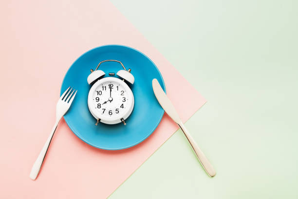 istock Intermittent fasting concept. White alarm clock on empty blue dish with knife and fork on pink-green background. top view, copy space. 1202142166
