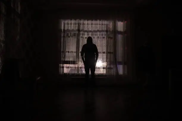 Silhouette of a man standing at a window inside the room. Dark mood conceptual image