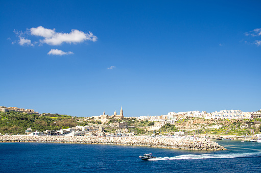 View of port village town Mgarr, catholic Ghajnsielem Parish Church and speedboat in the foreground on the Gozo island in front of blue sky with clouds, Malta