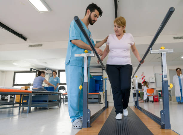 istock Female senior patient at physical therapy walking with the help of parallel bars and therapist next to her assisting 1202155246