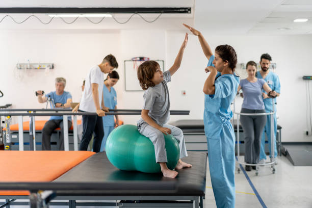 istock Latin american 7 year old boy during physical therapy sitting down on ball while following therapists arms at a physical recovery clinic 1202157454