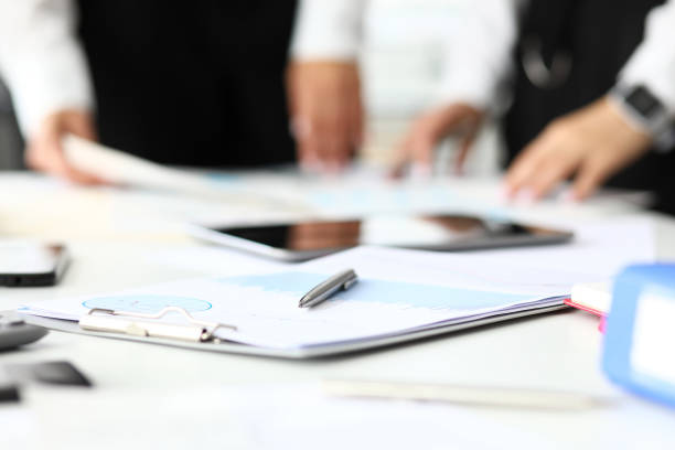 Silver pen lying at market index review paper graph clipped to pad with two workers standing in background close-up